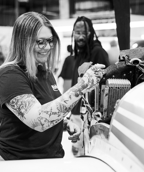 Smiling female aviation technician with tattoos and glasses working on an aircraft engine in a hangar, with a male colleague observing in the background.